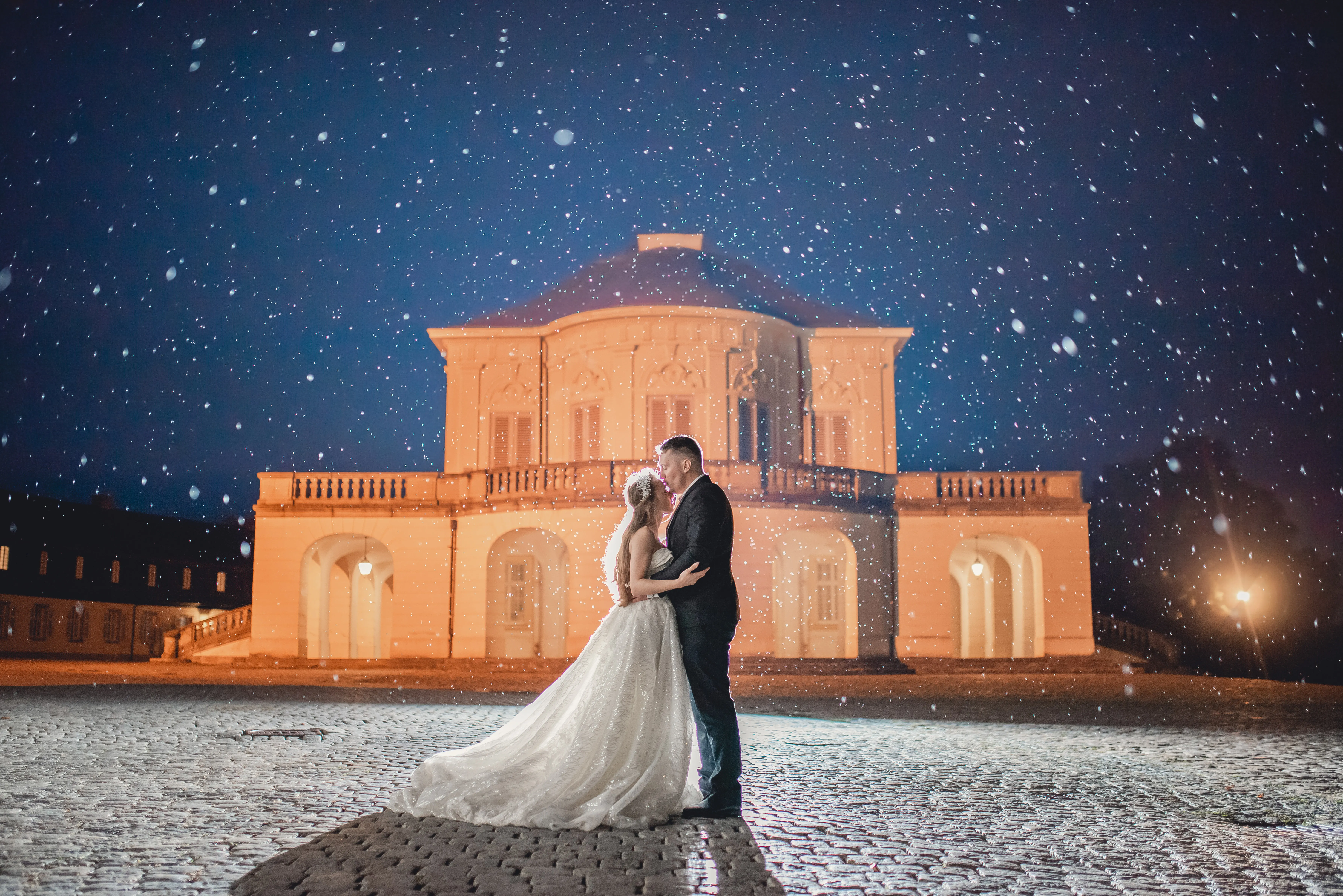 Brautpaar küsst sich nachts bei Schneefall vor einem beleuchteten Schloss
