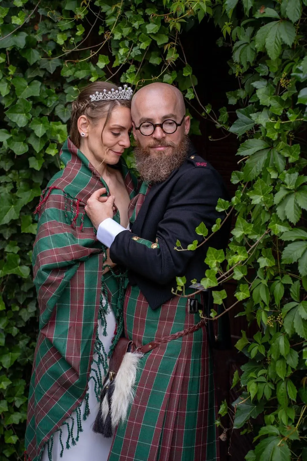 Braut in kariertem Tartan-Tuch umarmt ihren Bräutigam im Kilt vor einer Wand aus grünem Efeu