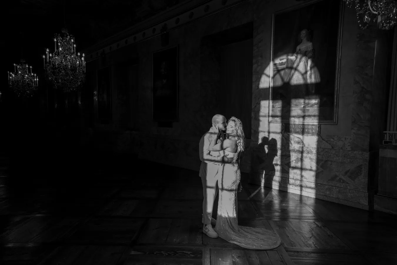 Schwarz-Weiß-Foto: Brautpaar umarmt sich in einem Festsaal mit Kronleuchtern, das Sonnenlicht durch ein Fenster wirft ihre Schatten an die Wand